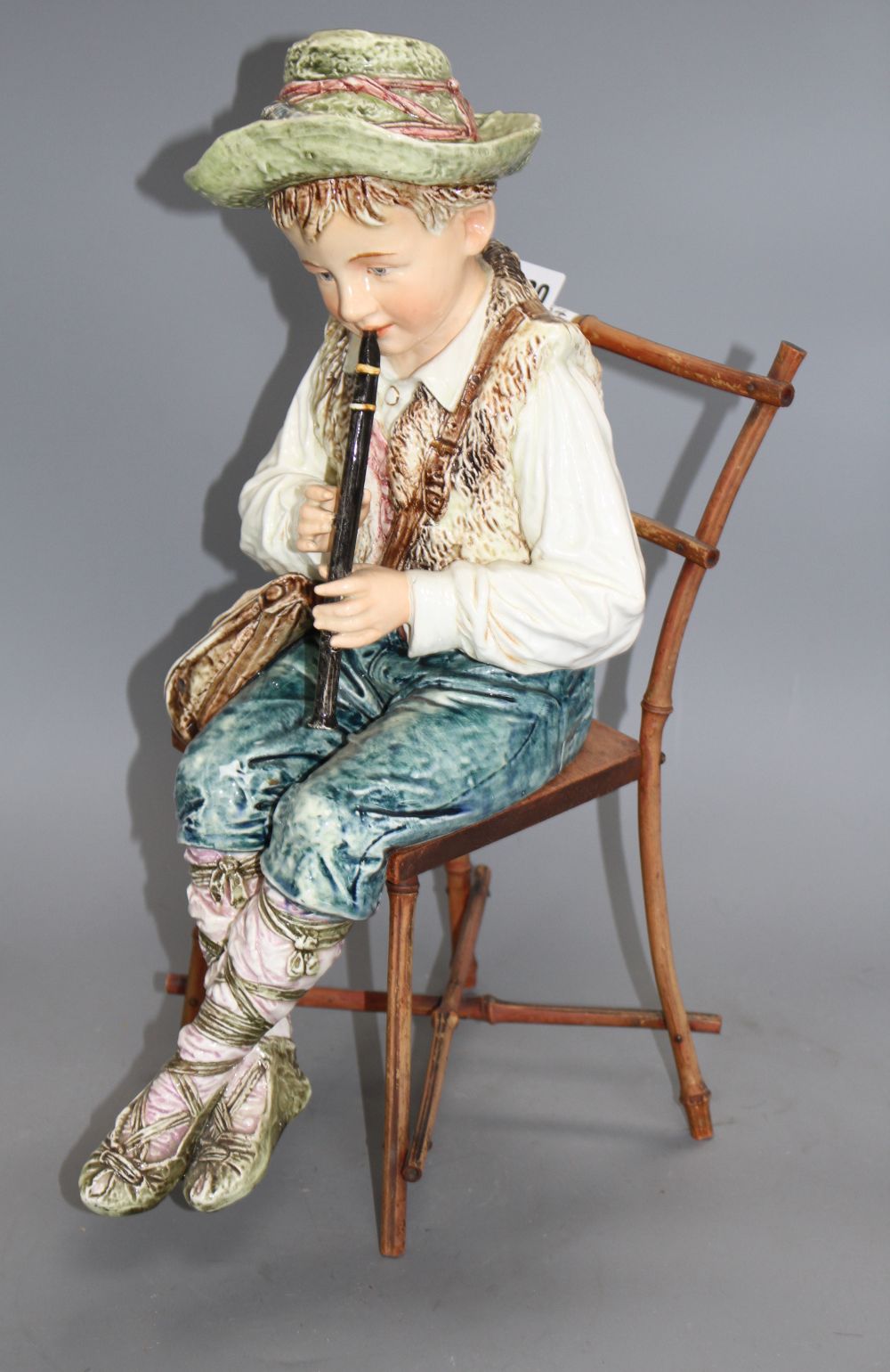 A late 19th century Austrian tin glaze pottery figure of a Tyrolean boy playing a flute, seated upon a bamboo wood chair, height 43cm